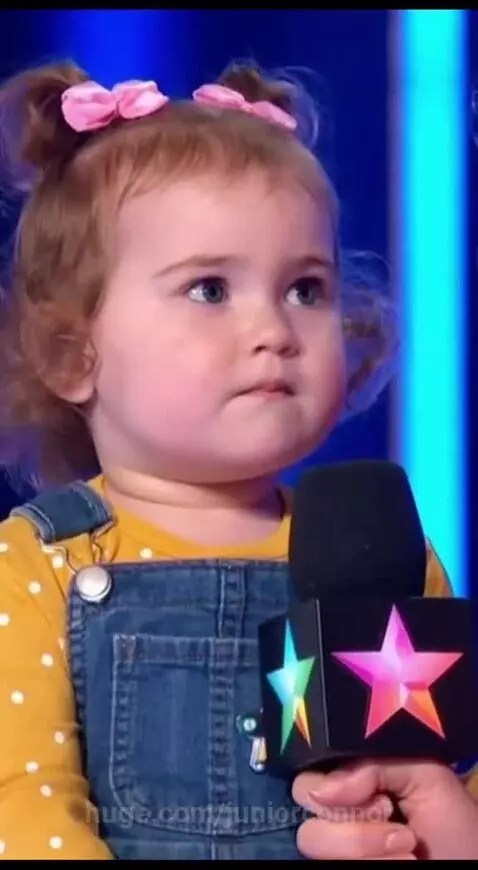 Red-haired baby with pigtails and pink bows, wearing a yellow polka dot shirt and denim overalls, sits on a stool.