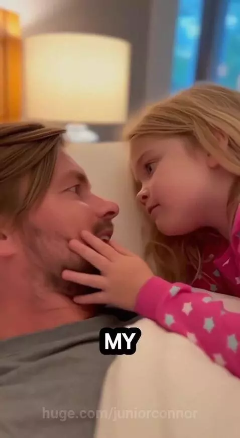 Blonde girl in pink pajamas with hand on dad's cheek, looking at him playfully.