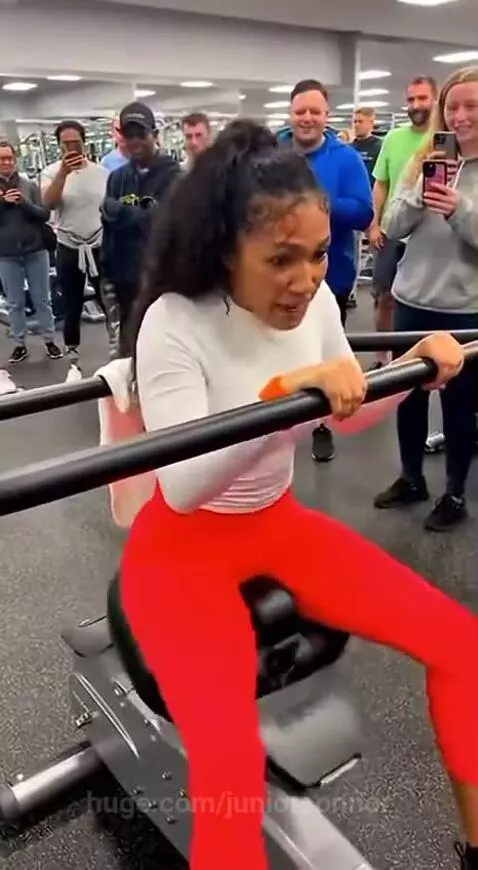 Woman with wide-eyed, scared expression on a vibrating rowing machine in a gym, surrounded by onlookers.