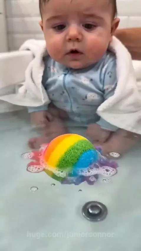 Baby with wide eyes watching a rainbow bath bomb fizz, then smiling at a rubber duck.