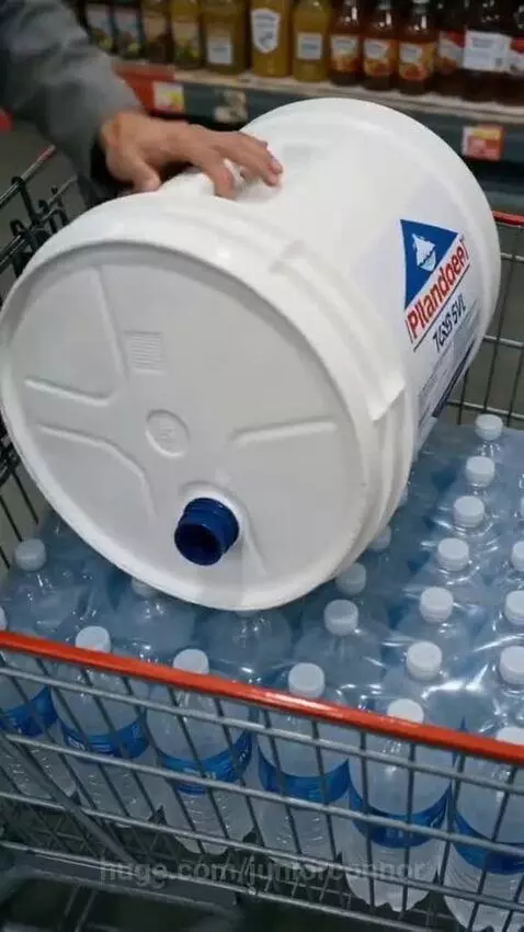 Man in grey jumpsuit pushing two full shopping carts in a store, carrying generator and water.