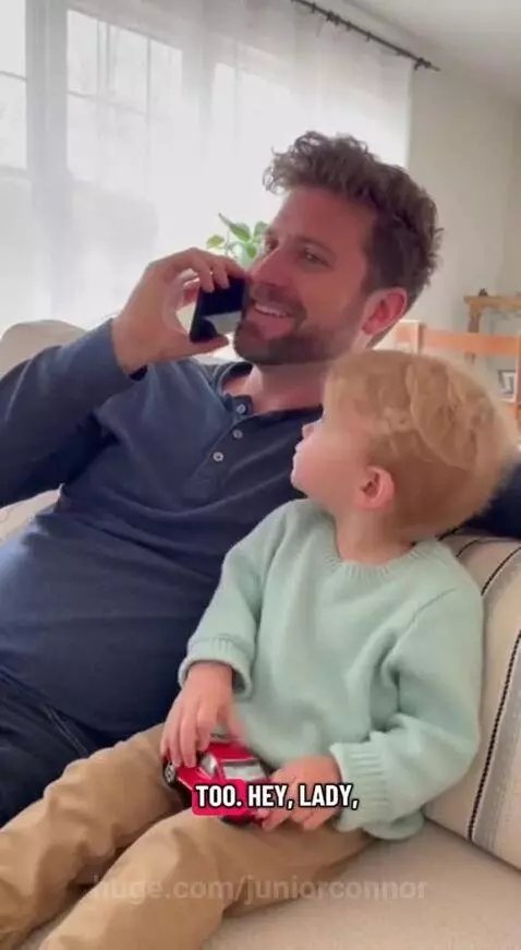 Young boy holding a toy car, looking sternly at a phone while his dad sits beside him.