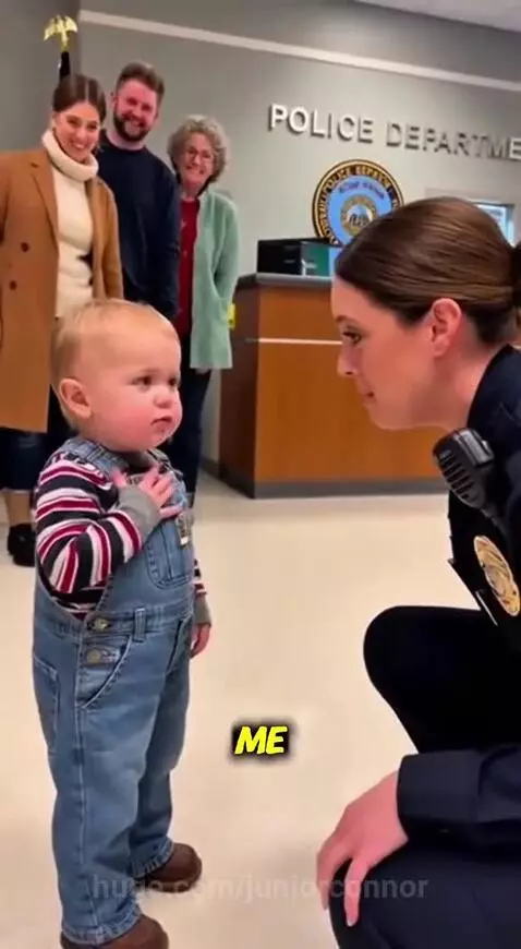 Toddler in overalls points at a kneeling female police officer, making her laugh with a sweet joke.
