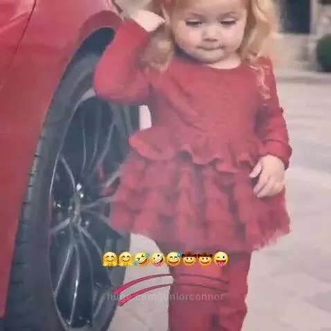 Young blonde girl in a red outfit playfully tapping a car tire with her hands and feet.