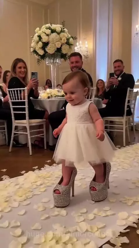 Baby girl in a white dress and oversized sparkly heels walks down a wedding aisle covered in rose petals.