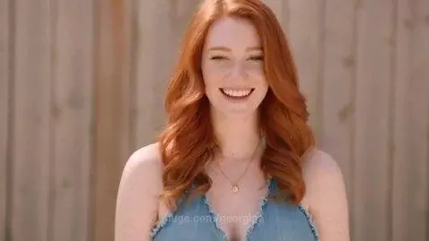 Red-haired woman with freckles smiling and laughing, wearing a denim halter top and short denim shorts, with a wooden fence background.