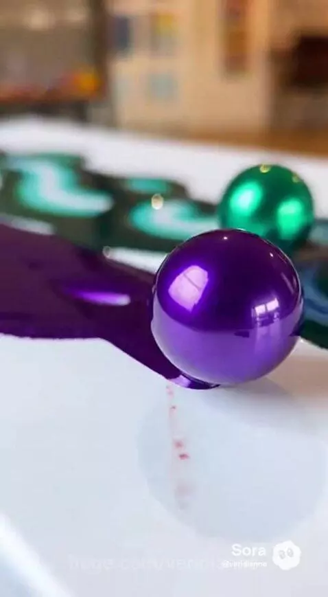 Close-up of purple and green paint swirls created by rolling metallic spheres on a white surface.