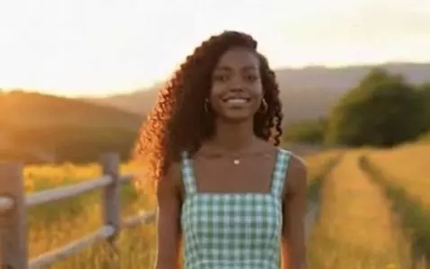 Woman with curly dark hair in a sundress walks through a sunlit grassy field at sunset.
