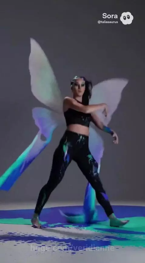 Woman with dark hair dancing in a studio with translucent butterfly wings and colorful paint splatters on her and the floor.