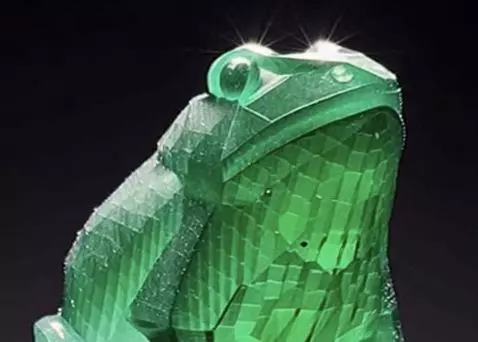 A faceted green emerald frog sculpture sitting on a dark reflective surface, catching the light.