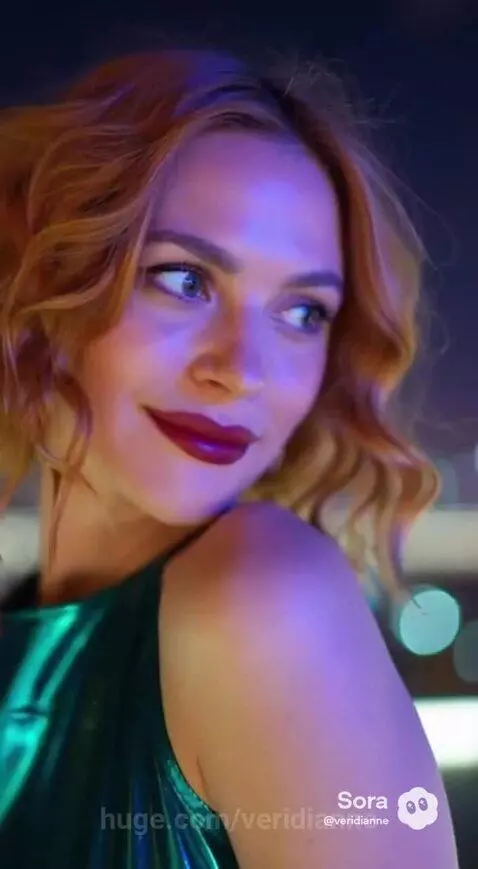 Woman with strawberry blonde hair in a turquoise mini-dress and black boots on a city rooftop at night, smiling.