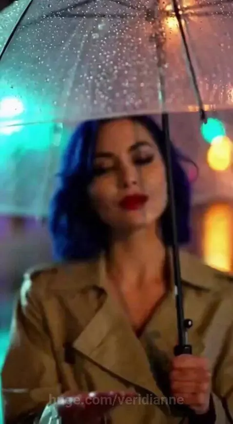 Woman with blue hair in a trench coat holding a striped umbrella on a wet city street at night.