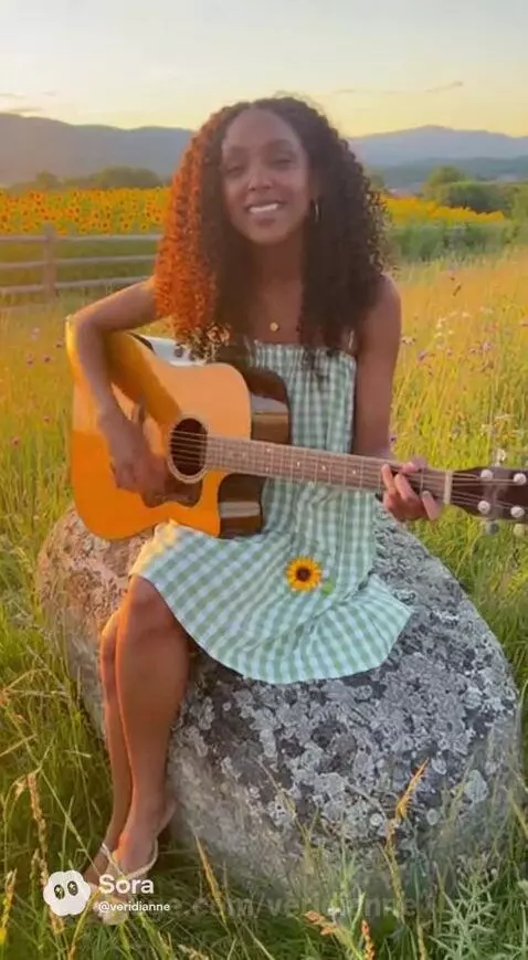 Woman with curly brown hair in a sunflower dress singing and playing guitar on a rock in a sunflower field.