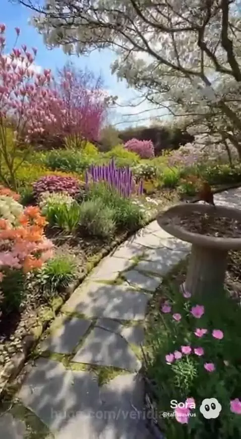 Stone path winding through a vibrant garden with colorful flowers, large trees, and a blue sky.