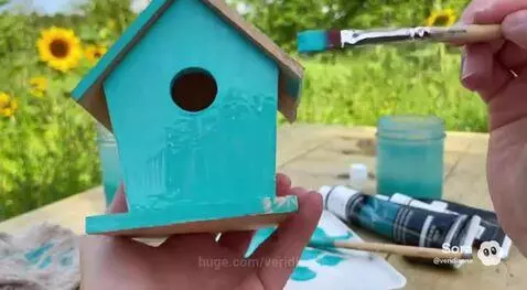 Close-up of hands painting a wooden birdhouse with bright turquoise paint, with sunflowers in the background.
