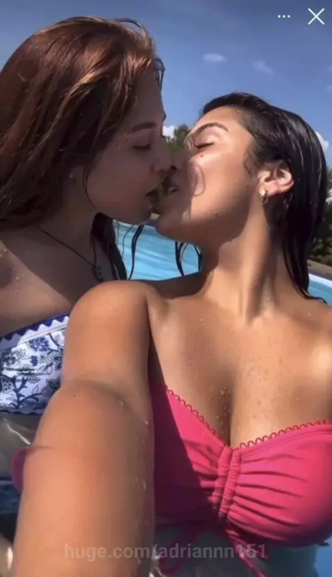Two women with smiles share an intimate kiss in a swimming pool on a sunny day, water droplets visible on their skin.