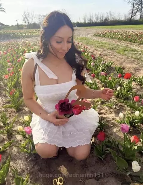 Woman with long dark hair in a white dress kneeling in a field of colorful tulips, holding a basket, with a beach in the background.
