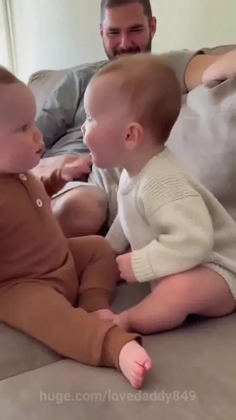 Two babies in outfits facing each other on a couch, making sounds, with a smiling dad in the background.