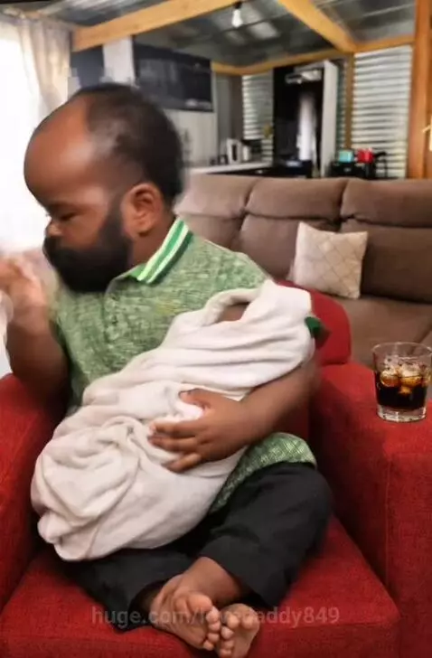 Man with dwarfism holding a swaddled baby, making playful expressions in a red armchair.