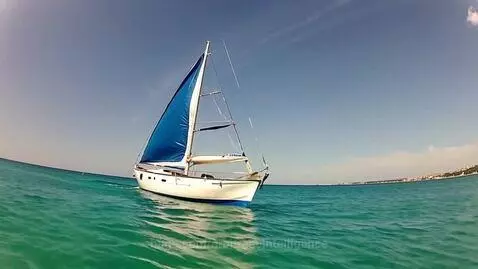 A white sailboat with a bright blue sail moving across calm turquoise ocean water under a clear blue sky.