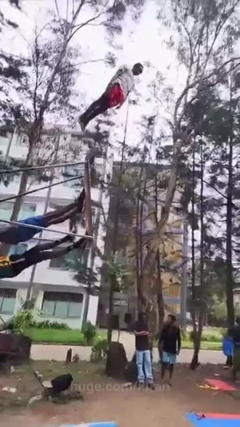 Man in red shorts and white shirt flips high in the air on a swing apparatus, landing on a cushion held by friends.