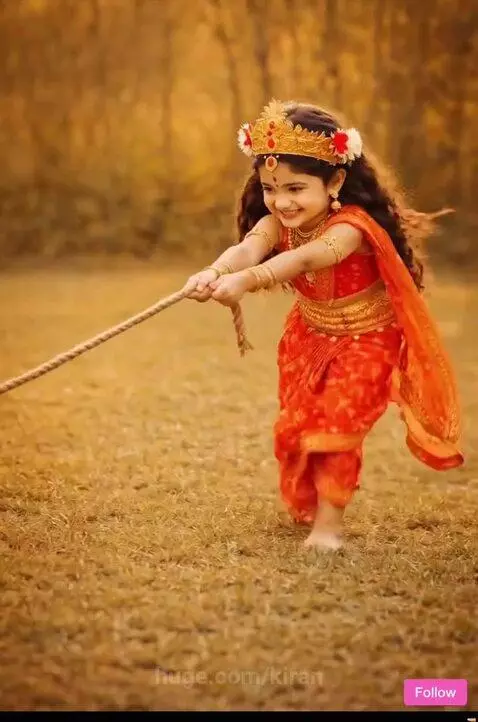 Young girl in traditional Indian attire and crown playing with a black goat in a grassy field.