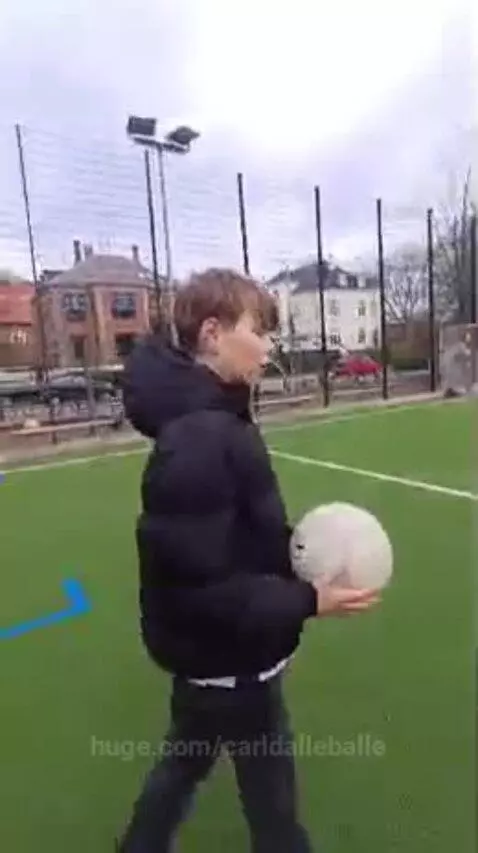 Young boy with light brown hair holding a white soccer ball on a green artificial turf soccer field.