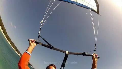 Person parasailing over a clear turquoise ocean with a white sandy beach and green vegetation visible below.