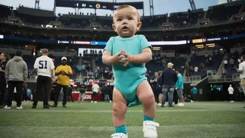 Baby in a blue onesie dancing on a football field during a stadium event.