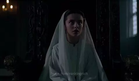 A young woman in a white habit sits on a dark chair, looking up with a serious expression.