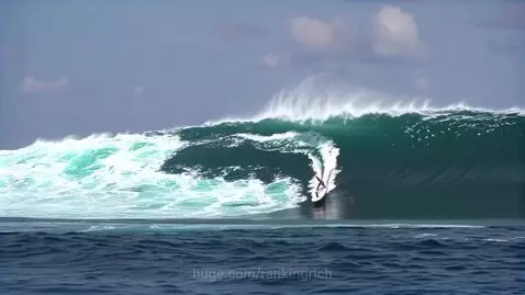 A surfer in a pink top rides inside the barrel of a huge, powerful blue-green ocean wave with a white crest.