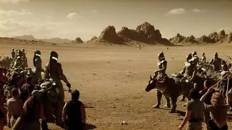 Aliens in futuristic armor riding dinosaurs charge at cavemen in a desert landscape.