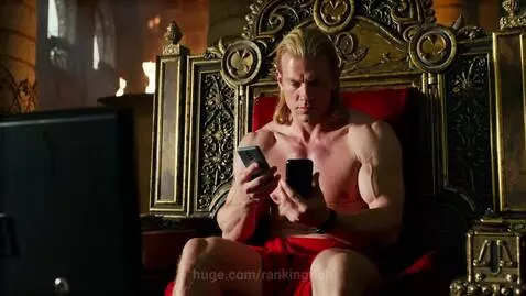 Muscular man with long blonde hair, dressed as a king, sitting shirtless on a golden throne and holding two smartphones.