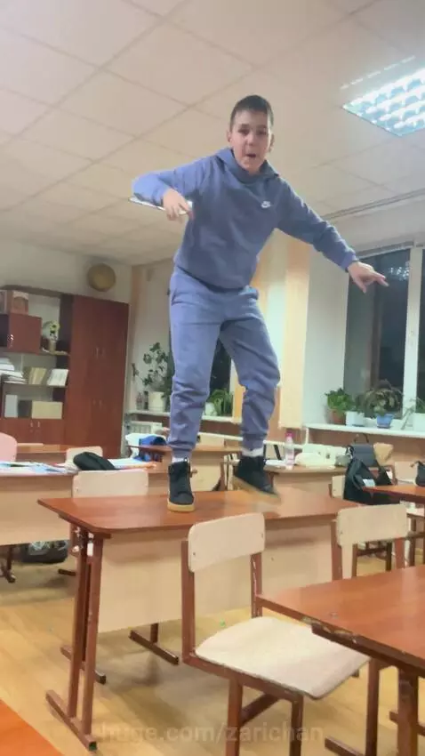 Young boy in purple tracksuit balancing on two wooden classroom desks, arms out for stability.