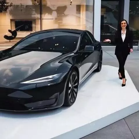 Woman in business suit smiling next to a sleek black Tesla on a white platform, modern building in background.