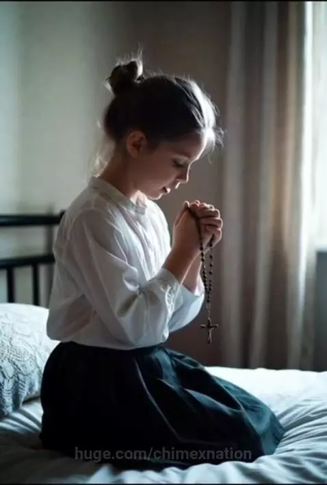 Young girl with hair in a bun, wearing a white shirt, kneels on a bed praying with a rosary.
