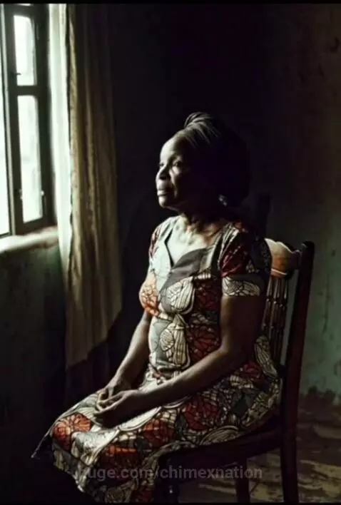 Elderly Black woman in a colorful dress and headwrap praying by a window.