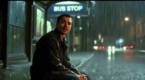 Man sitting alone at a rainy bus stop at night, looking dejected.