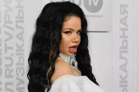 Close-up of a woman with dark wavy hair and glitter makeup spitting a thick, clear liquid from her mouth.