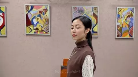 Asian woman with long dark hair in a ponytail, wearing a brown turtleneck vest over a white lace shirt, walks down a hallway with art.