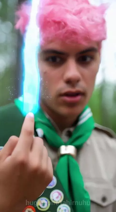 Young Eagle Scout with pink hair in uniform ignites a campfire with a blue flame from his finger.