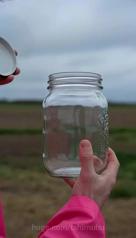 Person in pink raincoat holding a mason jar with a miniature tornado inside, with a large tornado in the background.
