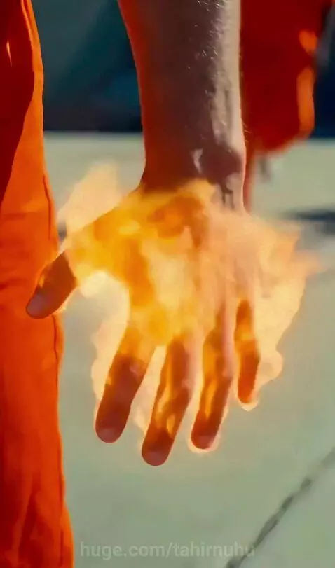 Man with pink hair in an orange jumpsuit ignites his hands with fire, punching an opponent, causing a large explosion in a prison yard.