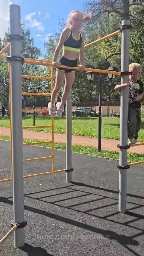 Young girl with braided hair performing acrobatic maneuvers on a yellow horizontal bar at an outdoor playground.