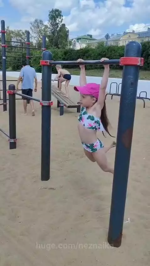 Young girl with ponytail doing pull-ups on a horizontal bar at a sandy outdoor gym on a sunny day.