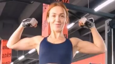 Woman with red braided hair in a sports bra and shorts doing assisted pull-ups in a gym.