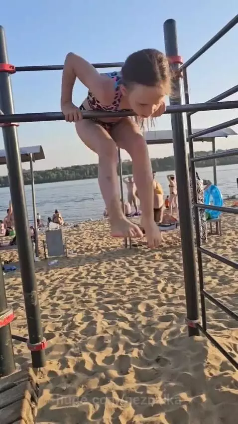 Young girl with long hair doing pull-ups on a beach bar, with the ocean and sunny sky in the background.