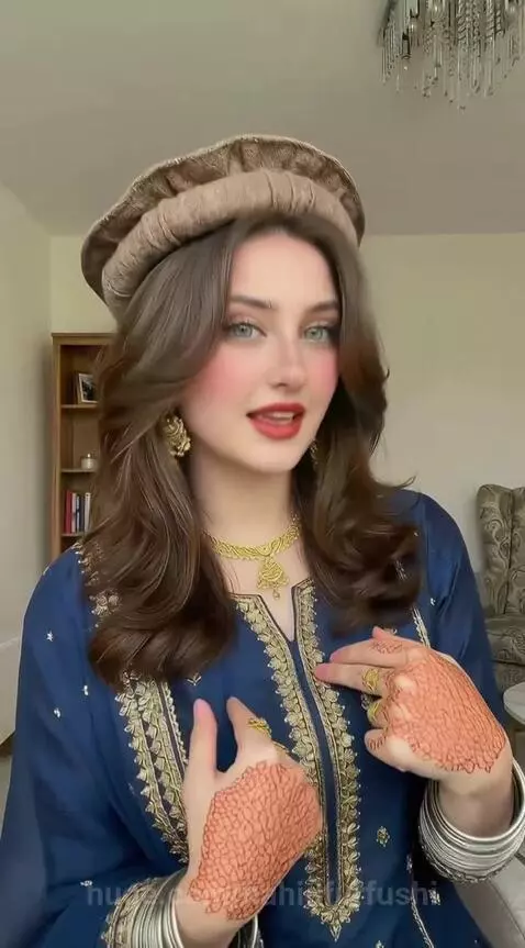 Young woman with long brown hair, blue eyes, wearing a blue embroidered dress, gold jewelry, and a hat, with henna on her hands.