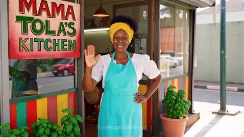 Woman in yellow headwrap and turquoise apron at Mama Tola's Kitchen storefront, waving hello.