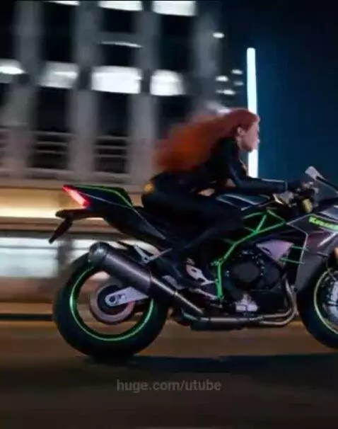 Woman with red hair in leather suit riding a black and green Kawasaki motorcycle through a city at night.